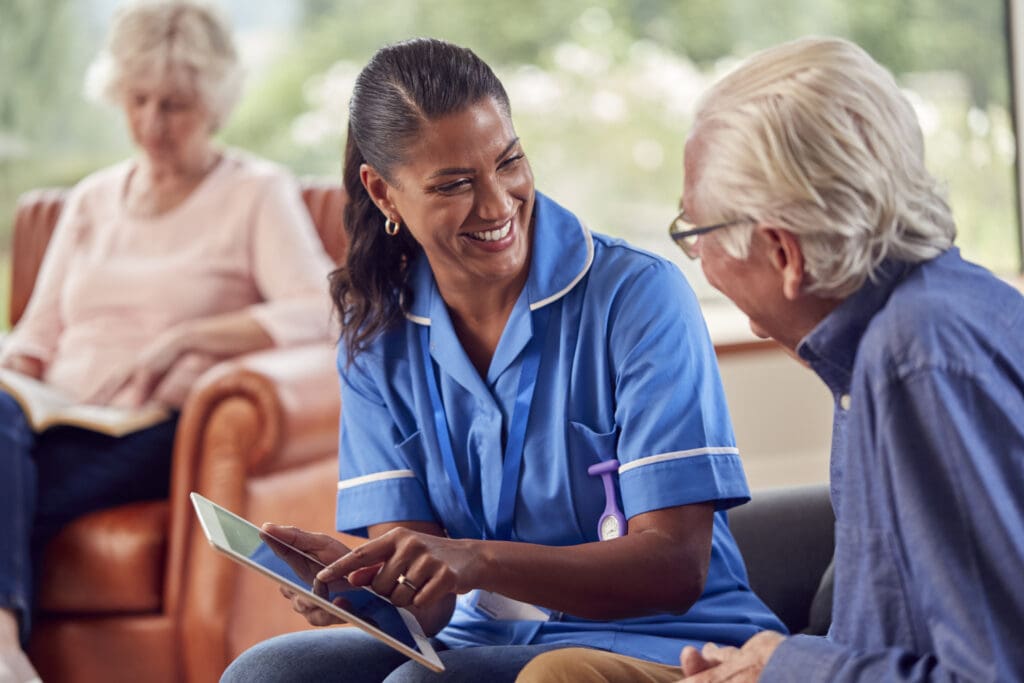 Senior Couple At Home With Man Talking To Female Nurse Or Care